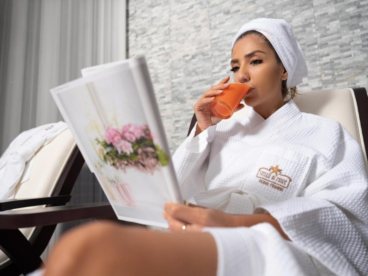 Stella Di Mare Dubai Marina Hotel Exterior foto The photo shows a woman sitting in a comfortable setting, likely a spa or relaxation area. She is wearing a white bathrobe and a towel on her head. In one hand, she holds a glass of orange juice, and in the other, she is reading a magazine or brochur